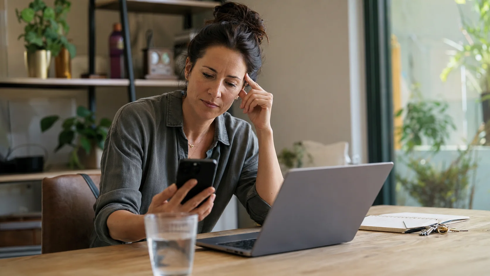A woman thoughtfully checking her phone beside a laptop during an account setup